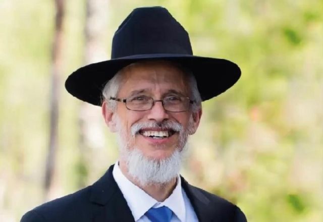 Rabbi Yisroel Meir Vogel, smiling, in a black hat and glasses, outdoors.